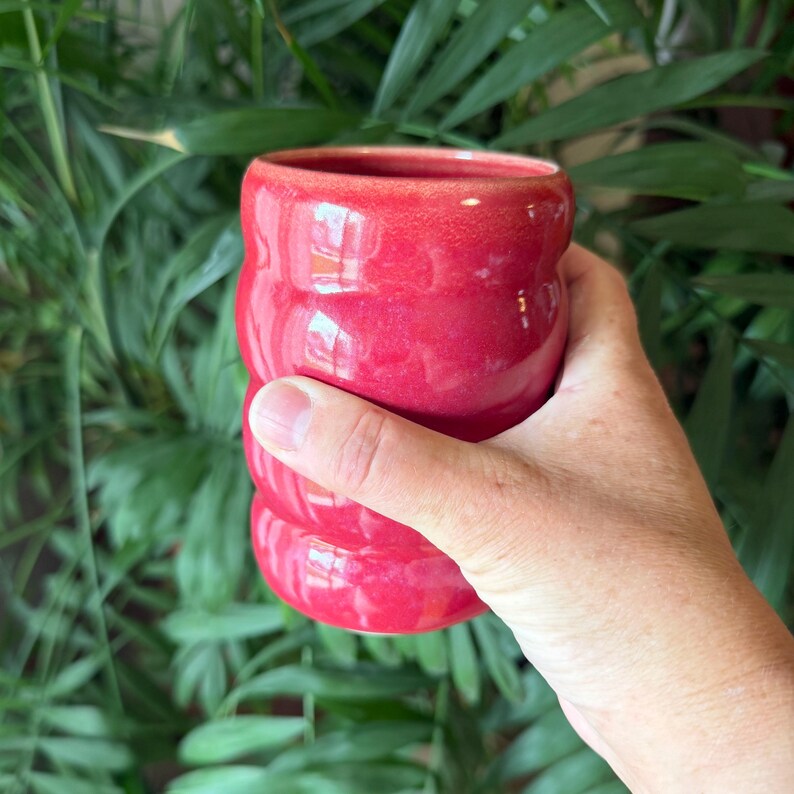 Puede incluir: Una mano sosteniendo un vaso de cer&aacute;mica rojo. El vaso tiene un dise&ntilde;o acanalado y un acabado brillante. El fondo es una vista borrosa de follaje verde.
