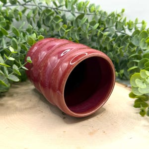 Puede incluir: Una taza cil&iacute;ndrica de cer&aacute;mica hecha a mano en un tono rojo intenso. La taza presenta un dise&ntilde;o acanalado en el exterior y un interior oscuro. La taza se coloca sobre un fondo de vegetaci&oacute;n y una superficie de madera.