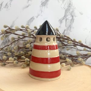 May include: A ceramic lighthouse figurine with a black top, tan body, and red stripes. The lighthouse has three small circular windows and is displayed on a wooden surface. Decorative branches with small, round buds are in the background.