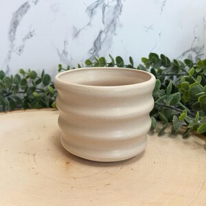 May include: A beige ceramic planter with a ribbed design. The small, round pot is unglazed and has a neutral color. The planter is suitable for small plants and succulents. The pot is approximately 4 inches in diameter and 3 inches tall.