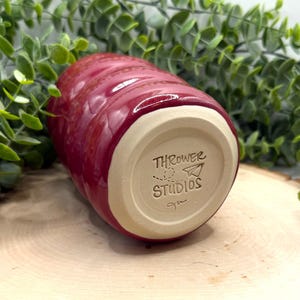 Puede incluir: Un vaso de cer&aacute;mica rojo hecho a mano con un exterior texturizado y acanalado. La base del vaso no est&aacute; esmaltada, revelando el color natural de la arcilla, y presenta el texto "THROWER STUDIOS" y un logotipo de avi&oacute;n de papel.