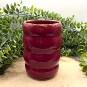 May include: A small, cylindrical, burgundy-colored ceramic tumbler. The handmade cup has a ridged design and a glossy finish. The background features green foliage and a wooden surface. This unique piece is suitable for beverages.