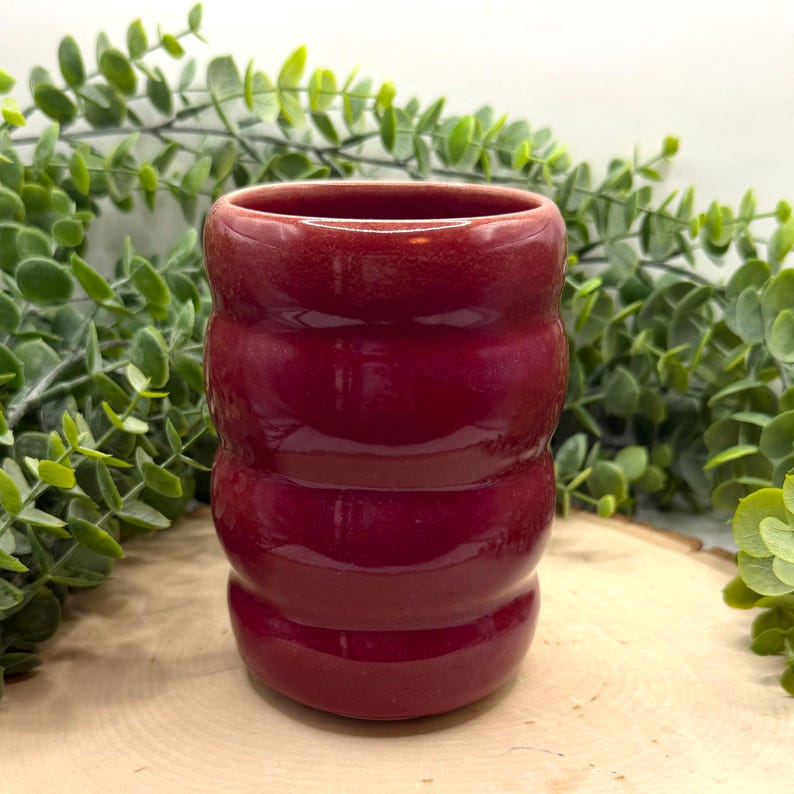 Puede incluir: Un peque&ntilde;o vaso cil&iacute;ndrico de cer&aacute;mica de color burdeos. La taza hecha a mano tiene un dise&ntilde;o acanalado y un acabado brillante. El fondo presenta follaje verde y una superficie de madera. Esta pieza &uacute;nica es adecuada para bebidas.