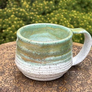 May include: Handmade ceramic mug with a rounded shape and a sturdy handle. The mug features a color gradient, transitioning from a speckled white base to bands of brown, green, and turquoise at the rim. The mug is sitting on a wooden surface.