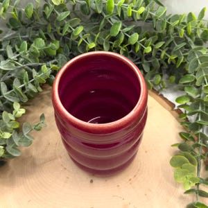 Puede incluir: Taza de cer&aacute;mica burdeos hecha a mano con un dise&ntilde;o acanalado. La taza cil&iacute;ndrica tiene una abertura ancha y un acabado brillante. La taza est&aacute; colocada sobre una superficie de madera, rodeada de follaje verde. Aproximadamente 10 cm de alto.