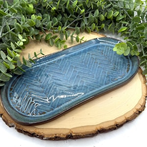 May include: A handmade, oval-shaped ceramic tray with a blue glaze and a textured herringbone pattern. The tray has a dark brown rim and is displayed on a wooden slice with green foliage in the background. A decorative piece for the home.