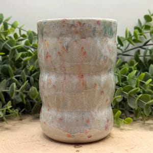 May include: A handmade ceramic tumbler with a speckled design. The off-white mug features a textured, ridged body and is adorned with colorful flecks of red, yellow, and blue. The rim has a subtle green hue, adding a touch of color to the design.