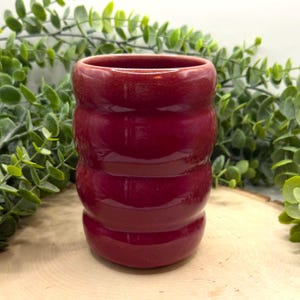 Puede incluir: Un jarr&oacute;n de cer&aacute;mica rojo oscuro con forma cil&iacute;ndrica y ranuras horizontales. El jarr&oacute;n tiene un acabado brillante y una abertura ancha. El fondo presenta follaje verde y una superficie de madera. El jarr&oacute;n mide unos 10 cm de alto.