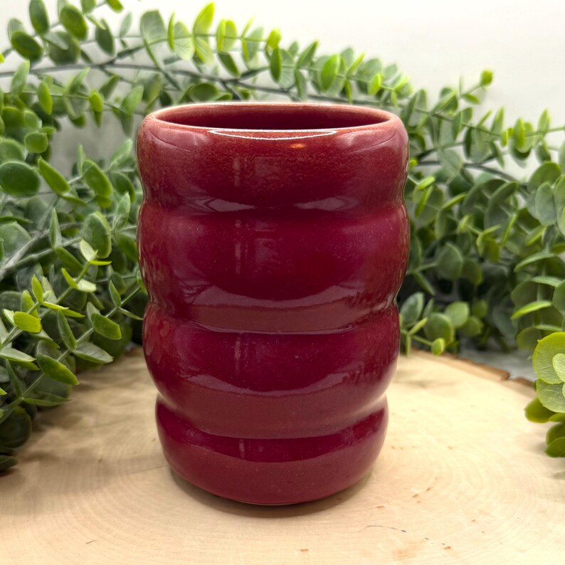 Puede incluir: Un jarr&oacute;n de cer&aacute;mica rojo oscuro con forma cil&iacute;ndrica y ranuras horizontales. El jarr&oacute;n tiene un acabado brillante y una abertura ancha. El fondo presenta follaje verde y una superficie de madera. El jarr&oacute;n mide unos 10 cm de alto.