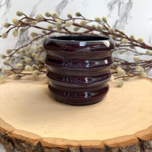 May include: A handcrafted, dark burgundy ceramic pot with a glossy finish. The pot features a ribbed design and a dark interior. It sits on a wooden slice, with a marble background and decorative branches.