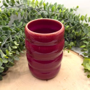 Puede incluir: Un vaso de cer&aacute;mica rojo oscuro brillante con un dise&ntilde;o acanalado. La taza cil&iacute;ndrica tiene una abertura ancha. El vaso se muestra sobre un fondo de follaje verde y una superficie de madera.