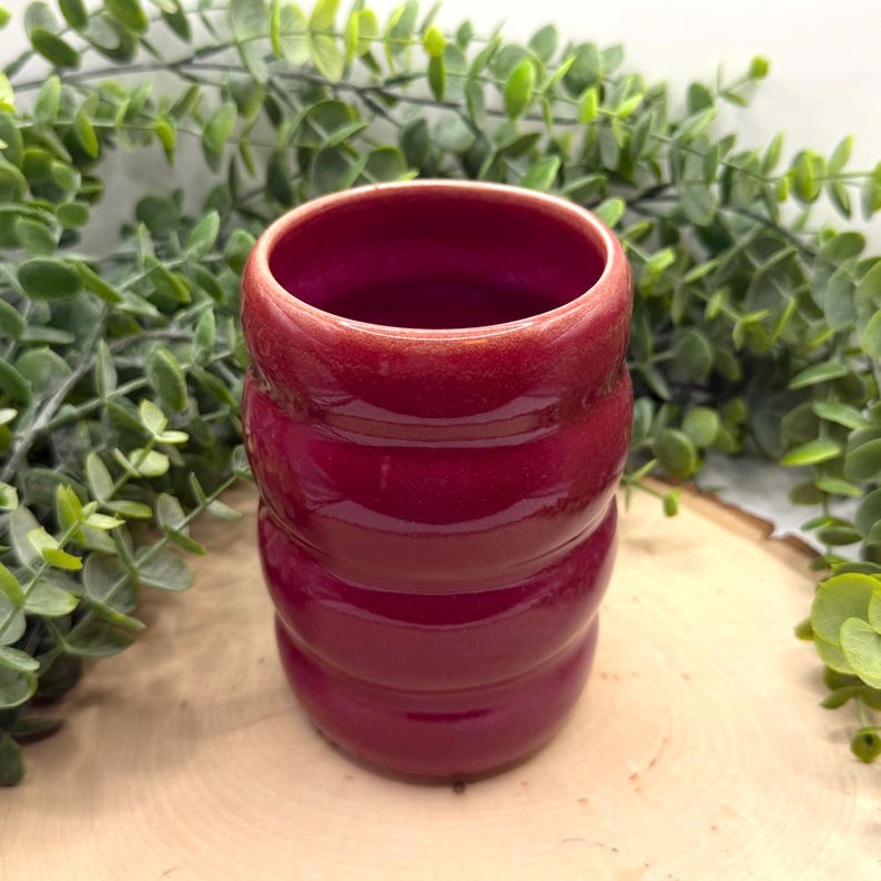 Puede incluir: Un vaso de cer&aacute;mica rojo oscuro brillante con un dise&ntilde;o acanalado. La taza cil&iacute;ndrica tiene una abertura ancha. El vaso se muestra sobre un fondo de follaje verde y una superficie de madera.