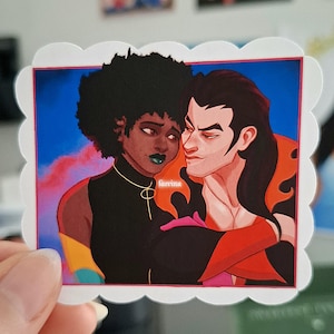 May include: A sticker with an illustration of two figures in an embrace. One figure has dark skin and an afro, wearing a black top, gold necklace, and green lipstick. The other has pale skin and long black hair, wearing a red and black outfit. The sticker has a white scalloped border.