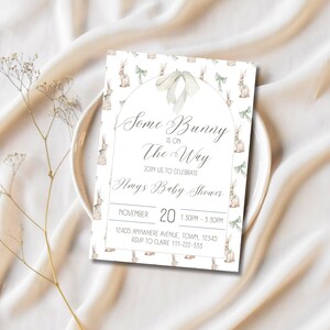 Op de afbeelding: Een witte babyshower-uitnodiging met een aquarel-illustratie van konijntjes en groene strikken. De tekst luidt "Some Bunny is on The Way" en bevat details over het evenement, inclusief de datum, tijd, locatie en RSVP-informatie.