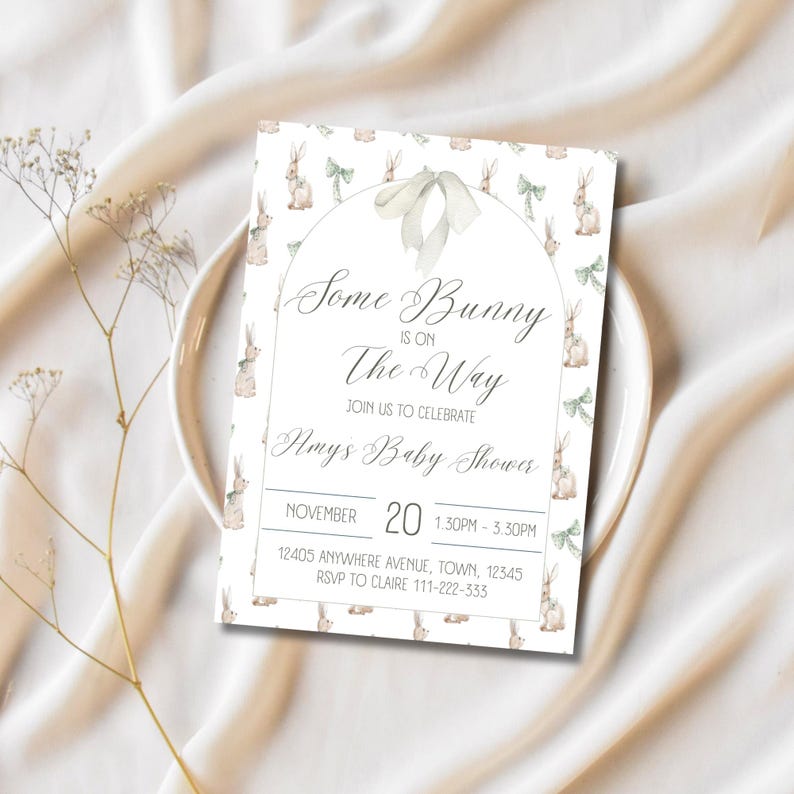 Op de afbeelding: Een witte babyshower-uitnodiging met een aquarel-illustratie van konijntjes en groene strikken. De tekst luidt "Some Bunny is on The Way" en bevat details over het evenement, inclusief de datum, tijd, locatie en RSVP-informatie.