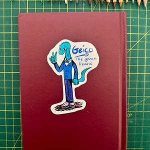 Op de afbeelding: Een bordeauxrood boek met een sticker met een cartoonhagedis in een pak, met de tekst "Geico The green lizard". Gekleurde potloden zijn langs de bovenrand van het boek gerangschikt, op een groene snijmat.