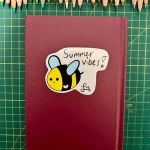 May include: A burgundy-colored book with a sticker featuring a cartoon bee and the words "Summer vibes!". A collection of colored pencils in various shades are arranged at the top of the image.