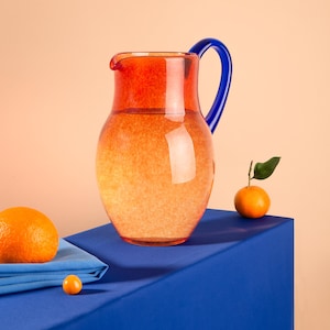 Op de afbeelding: Een rode en blauwe glazen kan gevuld met water staat op een blauwe tafelkleed met twee sinaasappelen en een klein oranje vruchtje ernaast.