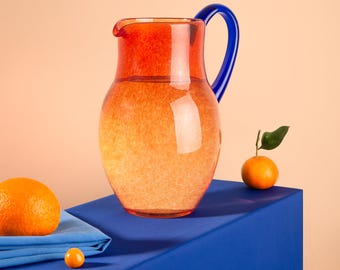 May include: A red and blue glass pitcher filled with water sits on a blue tablecloth with two oranges and a small orange fruit beside it.