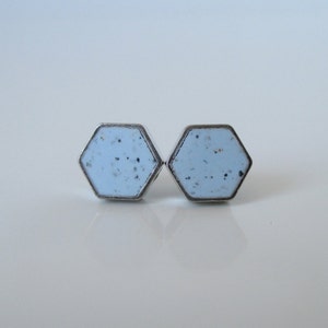 Puede incluir: Un par de pendientes de botón hexagonales. Cada pendiente presenta un centro de esmalte azul claro con motas oscuras, engastado en un marco de metal plateado. Los pendientes se muestran sobre una superficie blanca.
