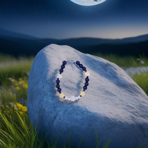 Peut inclure: Un bracelet de perles avec des perles bleu marine, blanches et jaunes. Le bracelet a un mélange de perles rondes et en forme d'étoile, avec un fermoir argenté. Le bracelet est présenté sur un rocher gris avec un fond de ciel nocturne.