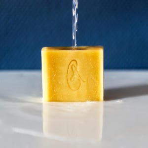 May include: A yellow bar of soap with a stylized ear design on the front. The soap is being rinsed with water.