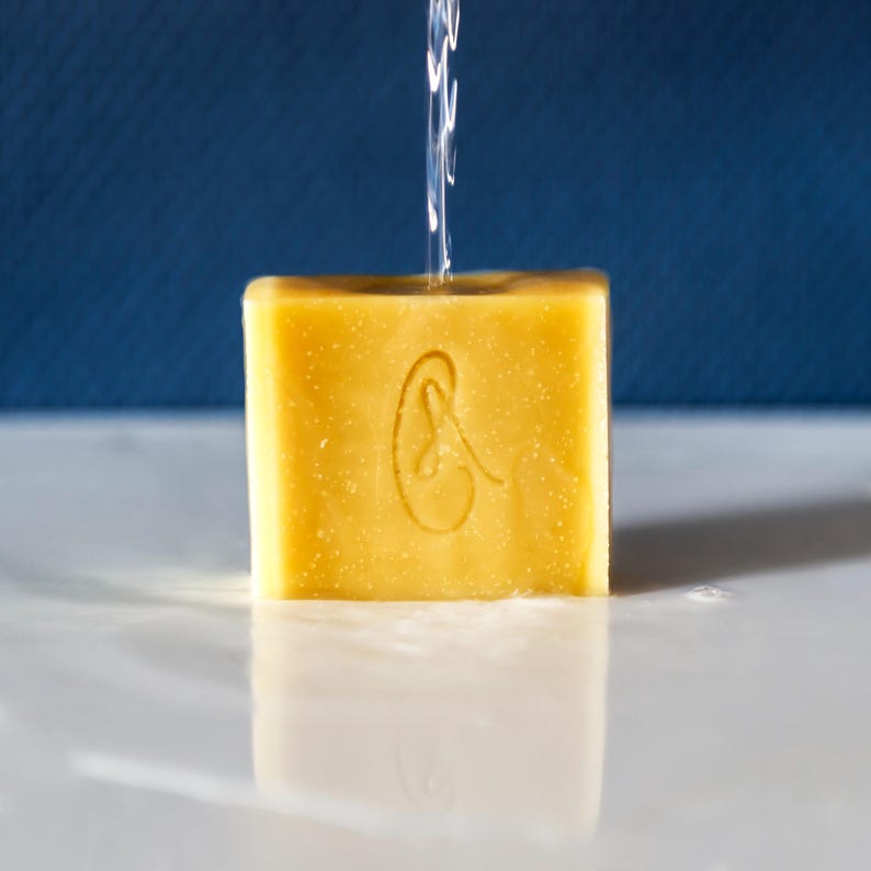 May include: A yellow bar of soap with a stylized ear design on the front. The soap is being rinsed with water.