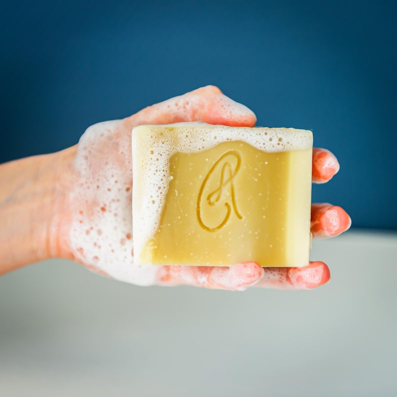 May include: A hand holding a bar of soap with a letter "A" embossed on it. The soap is surrounded by white bubbles.