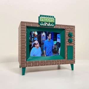 Może przedstawiać: Miniaturowy model telewizora z zewnętrzną powierzchnią przypominającą cegłę i zielonymi akcentami. Ekran wyświetla scenę z dwiema osobami. Na górze modelu znajduje się zielony znak z napisem "PADDYS". Telewizor ma zielone pokrętła i zielony wzór przypominający głośnik z boku.