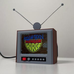 Könnte beinhalten: Ein braun-graues, fernsehförmiges Objekt mit zwei dünnen Metallantennen an der Oberseite. Der Bildschirm zeigt den Text Aqua Teen Hunger Force vor einem Ziegelmauerhintergrund. Die Vorderseite hat einen roten Knopf, einen Drehregler und vier kleine graue Tasten.