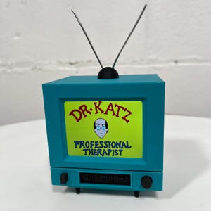 May include: A teal blue television with a black antenna and a screen displaying the text "DR. KATZ PROFESSIONAL THERAPIST" in yellow and red lettering.