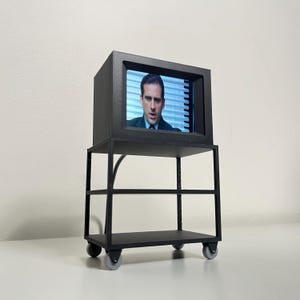May include: A black miniature television set with a person on the screen, mounted on a matching black cart with two shelves and four wheels. The person on the screen is wearing a suit and tie.