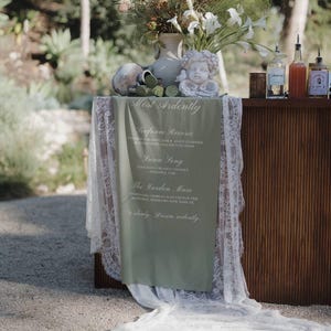 May include: A drinks bar with a green menu listing cocktails, draped over the front. The bar is decorated with flowers, a cherub statue, and bottles. The background is a blurred outdoor scene with trees.
