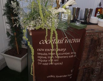 Pode incluir: Um menu de cocktails castanho com texto branco, apresentando os ingredientes das bebidas. O menu está drapeado sobre um bar de madeira, adornado com arranjos florais e ferramentas para fazer cocktails. Velas altas e garrafas também são visíveis.