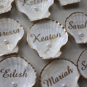 Puede incluir: Primer plano de varias tarjetas de lugar de concha blanca con borde dorado y nombres caligráficos. Cada concha tiene una pequeña perla en el centro. Los nombres están escritos en una fuente dorada. Ideal para bodas o eventos especiales.