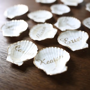 Könnte beinhalten: Weiße Muschelkarten mit goldenen Rändern und personalisierten Namen in goldener Schrift. Diese dekorativen Muscheln sind auf einer Holzoberfläche angeordnet, ideal für eine Hochzeit am Strand. Die Namen "Erin", "Keaton", "Carissa" und "Alexa" sind sichtbar.