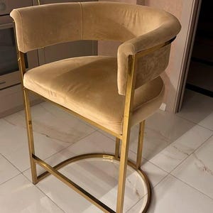 May include: A gold metal bar stool with a beige velvet seat. The stool has a curved back and a simple, modern design.
