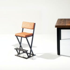 Könnte beinhalten: Ein brauner Leder-Barhocker mit einem schwarzen Metallgestell. Der Hocker hat eine Fußstütze und ein schlichtes, modernes Design.