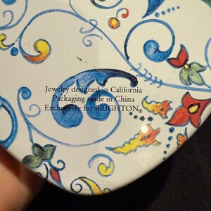 Puede incluir: Lata blanca en forma de coraz&oacute;n con un dise&ntilde;o floral azul y amarillo. El texto en la lata dice "Jewelry designed in California Packaging made in China Exclusively for BRIGHTON."