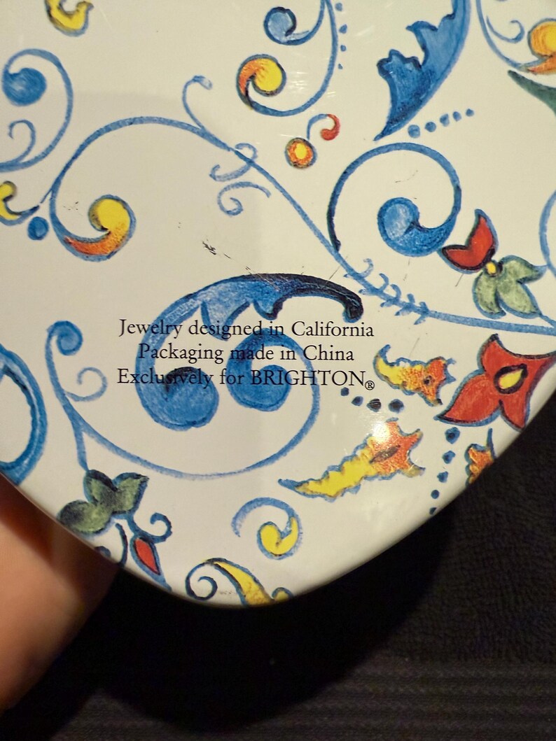 Puede incluir: Lata blanca en forma de coraz&oacute;n con un dise&ntilde;o floral azul y amarillo. El texto en la lata dice "Jewelry designed in California Packaging made in China Exclusively for BRIGHTON."