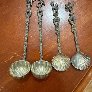 May include: Four antique silver spoons with ornate handles and decorative bowls. Two spoons have round bowls, while the other two have shell-shaped bowls. The handles feature detailed figures and floral designs.