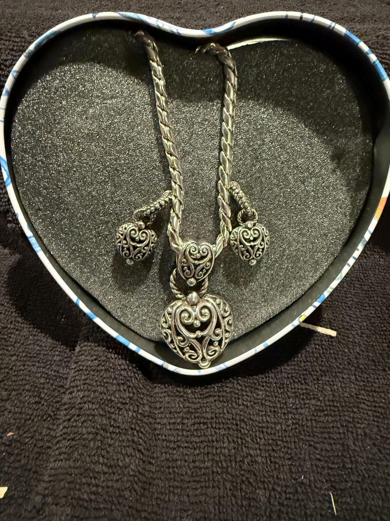 Puede incluir: Un collar de plata en forma de coraz&oacute;n con dos peque&ntilde;os colgantes en forma de coraz&oacute;n que cuelgan de la cadena. El collar est&aacute; en una caja en forma de coraz&oacute;n.