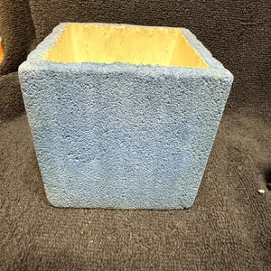 Peut inclure: Un pot de fleurs carré en matériau rugueux et texturé. Le pot est peint en bleu clair avec un intérieur jaune.