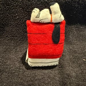 May include: A plush toy of Snoopy, the beagle from the Peanuts comic strip, sleeping in his red doghouse. The doghouse has a white trim and a black door.