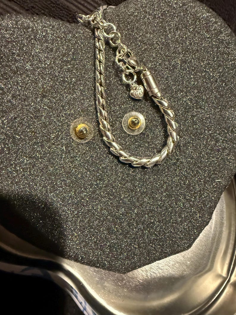 Puede incluir: Un brazalete de plata con un dije en forma de coraz&oacute;n y un cierre magn&eacute;tico. El brazalete se muestra sobre un fondo negro.