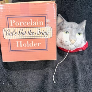 May include: A porcelain cat-shaped string holder with gray and white fur, green eyes, and a red collar. A piece of white string extends from the cat's mouth. Next to it is a cardboard box labeled Porcelain Cat's Got the String Holder.