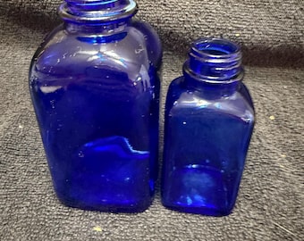 Peut inclure: Deux bouteilles en verre bleu cobalt de tailles différentes. Les bouteilles ont une forme arrondie, légèrement irrégulière, avec un col étroit et un bord fileté. Les bouteilles sont vides et semblent être vintage.