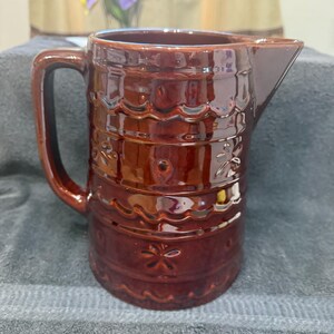 Könnte beinhalten: Ein brauner Keramik-Krug mit einem dekorativen Design. Der Krug hat eine breite Öffnung und einen Henkel. Das Design zeigt ein sich wiederholendes Muster aus gewellten Rändern und floralen Motiven.