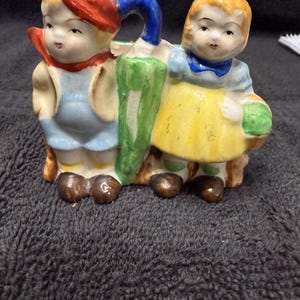 May include: A vintage ceramic planter shaped like two children holding an umbrella. The boy is wearing a red hat and blue overalls. The girl is wearing a yellow dress and a blue bow.