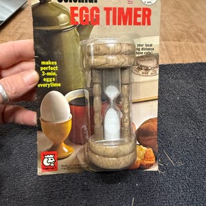 May include: A wooden egg timer with a clear glass hourglass inside. The timer is packaged on a card with the text "Colonial Egg Timer" and "makes perfect 3-min. eggs everytime".